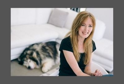 Image shows Rachel Amphlett posed in a studio laughing at the camera while the photographer's dog is sleeping on the floor behind her with its tongue out