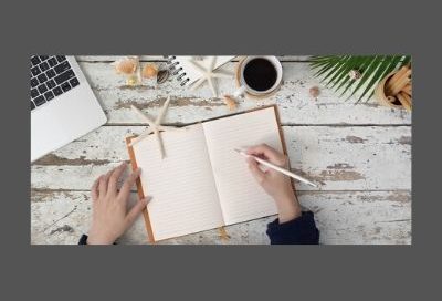 Image shows an overhead view of a woman holding a pencil above a ruled notebook with a laptop computer and a cup of black coffee next to her