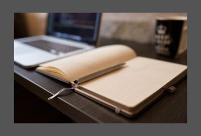 Images shows an open ruled notebook beside a laptop computer with a pen resting in the crease of the notebook