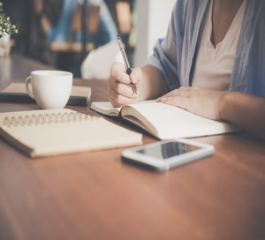 Image shows a woman writing in a notebook at a desk with a smartphone and a cup beside her