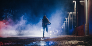 A man in a hooded sweatshirt running away from a car's headlights at night in an industrial area of the city